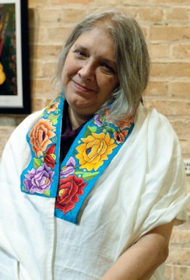 (ID: A photo of a smiling woman with short gray hair. She is wearing a cream-colored wrap that has a multicolored floral border.)