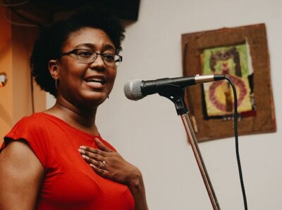 (ID: Camisha Jones stands in front of a microphone reciting a poem. She wears glasses and a sleeveless red top. She has brown skin and natural hair.)