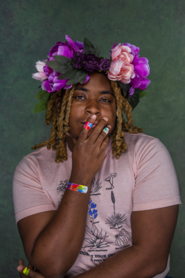 ID: KB — a Black person with brown skin — has a green background. They are wearing a flower crown, with pink and purple flowers, along with green leaves. Their hair is braided and blond. Their hand is slightly covering their mouth, and both their arms and hands have band-aids on them. KB is wearing a pink shirt with black and blue images of cactus plants on them. The elbow of one arm rests on their other arm. They are looking directly at the camera.